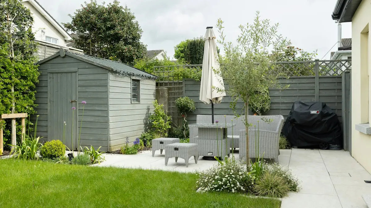 A grey timber apex shed in the corner of a back garden, with room to walk round it beside the fence and patio.