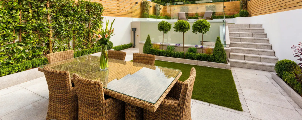 A wicker dining set on a pale patio in a landscaped back garden with a lawn and steps.